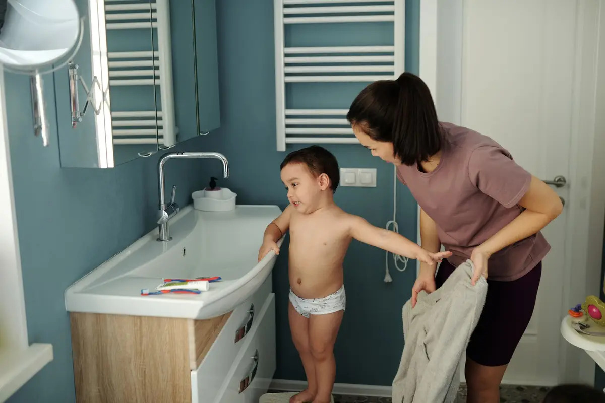 家長在浴室協助幼兒練習盥洗，孩子穿著學習褲站在洗手台前（示意圖）