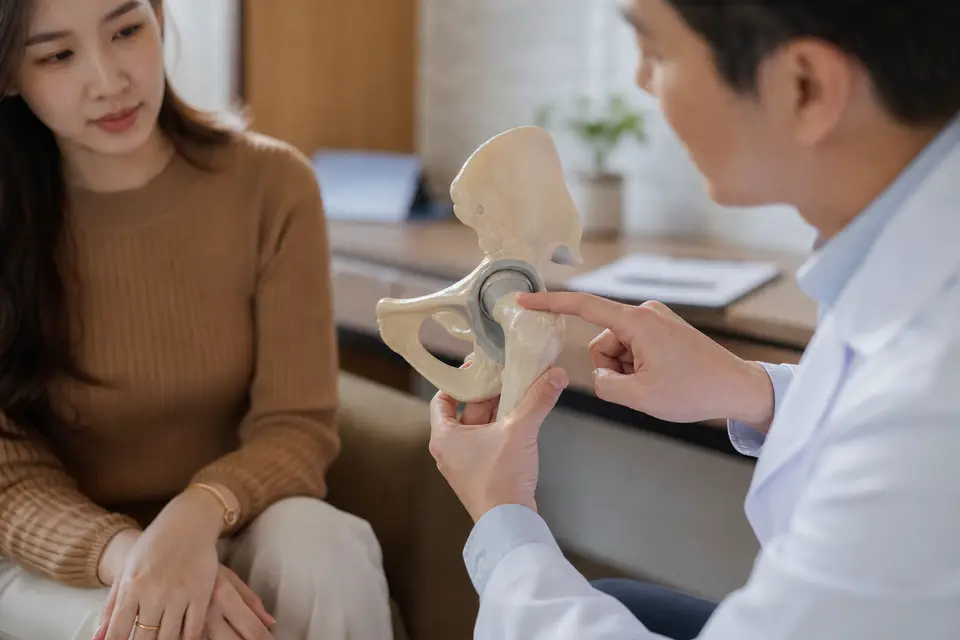 醫師在診間拿著髖關節骨骼模型，向坐著的患者說明疼痛位置（示意圖）