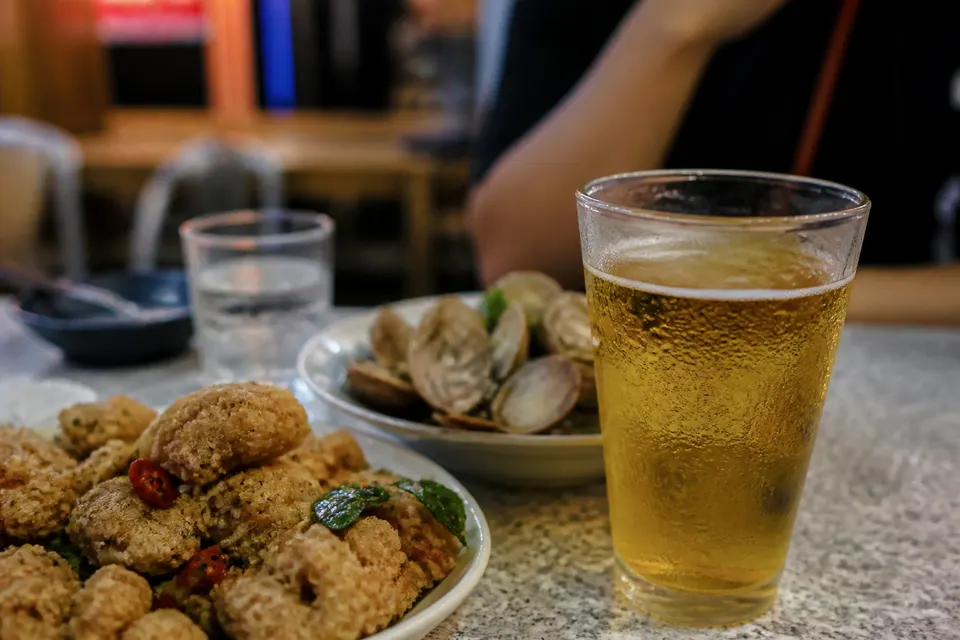 餐桌上一杯冒著水珠的冰啤酒旁邊放著一盤鹽酥雞與蛤蜊，背景有一杯白開水